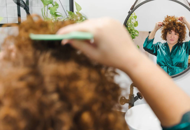 A chubby female with curly ginger hair brushing her hair in a bathroom mirror - Fotoğraf, Görsel