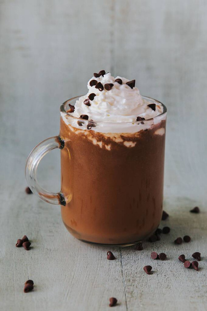 A vertical closeup of a glass of hot chocolate on a table, with cream on top and chocolate pieces on the side - Foto, Bild