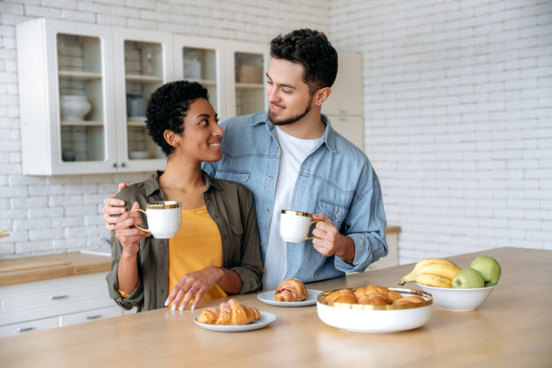 Happy husband and wife of different nationalities have tasty weekend breakfast in the kitchen at home with croissants and coffee, enjoying spend time together, looks at each other with love, smile - Photo, Image