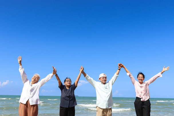 Group of four cheerful elderly old senior friends travel outdoor together, holding hand in line and raising up on beach blue sky, enjoy spending time, resting and relaxing on holiday vacation. - Photo, Image