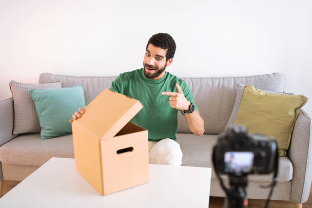 Positive junge Blogger aus dem Mittleren Osten zeigen mit dem Finger auf auspackende Pappkartons und drehen Video vor laufender Kamera im Wohnzimmerinneren. Blog-Empfehlung, Online-Shopping-Review, Lieferung nach Hause - Foto, Bild