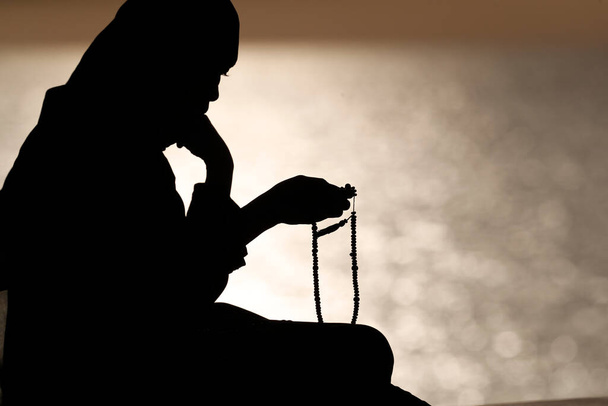 Siluetti musliminaisesta pitämässä rukoushelmiä käsissään ja rukoilemassa auringonlaskun aikaan. Uskonnon rukoilemisen käsite - Valokuva, kuva