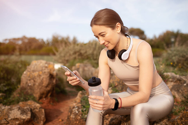 Positive young caucasian lady in sportswear, wireless headphones resting, typing on smartphone, hold bottle of water. Chat, app for body care, weight loss and blog for workout outdoor - Photo, Image