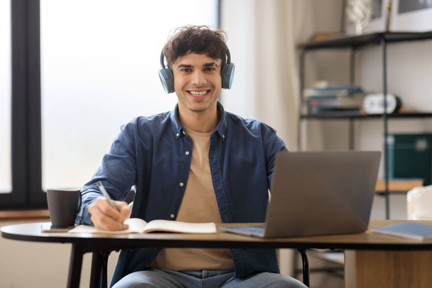 Vidám arab férfi laptoppal és jegyzetel a kamerába, fülhallgatót visel, ül az asztalnál a modern irodában. Portré a sikeres diák srác tanulás Online. Elektronikus tanulás - Fotó, kép