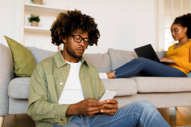 Poważny czarny człowiek Websurfing za pomocą smartfona, podczas gdy jego żona przeglądanie Internetu na komputerze w domu. Para komunikujących się i pracujących online. Styl życia ludzi i technologii. Skupienie selektywne - Zdjęcie, obraz