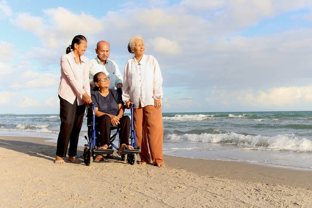 Group of four cheerful elderly old senior have a trip outdoor together, happy disabled senior elderly woman in wheelchair travel with friends on beach, enjoy spending time on vacation holiday. - Photo, Image