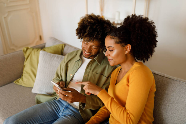 Cheerful African American Wife Pointing At Smartphone Browsing Internet Together With Husband Sitting On Sofa At Home. Couple Using Phone And Advertising New Mobile Application. Gadgets Concept - Foto, Imagen