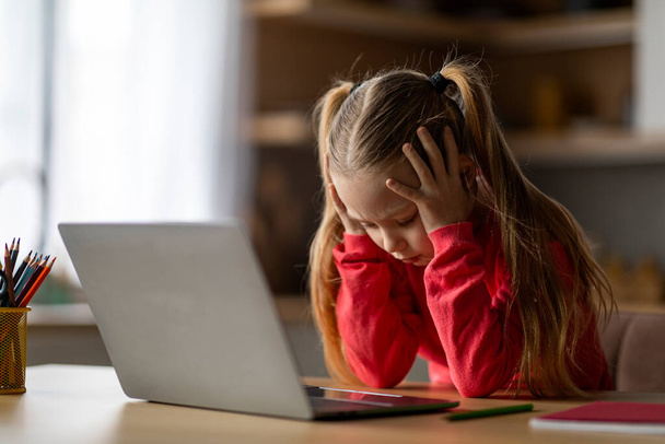 Schwierigkeiten bei der Einschulung. Verärgertes kleines Mädchen, das zu Hause einen Laptop benutzt und den Kopf berührt, gestresste Frühchen, die es satt haben, Hausaufgaben zu machen, depressives Kind, das am Schreibtisch mit Computer sitzt, freier Platz - Foto, Bild