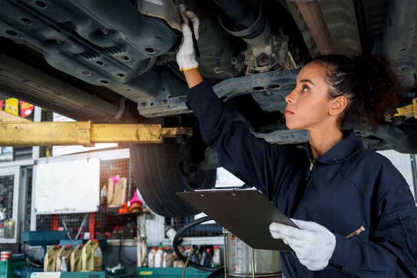 Beautiful female auto mechanic checking wheel tires in garage, car service technician woman repairing customer car at automobile service, inspecting vehicle underbody and suspension engine system. - Zdjęcie, obraz