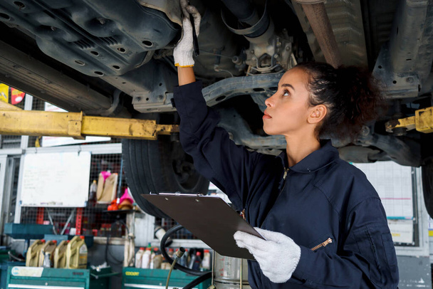 Beautiful female auto mechanic checking wheel tires in garage, car service technician woman repairing customer car at automobile service, inspecting vehicle underbody and suspension engine system. - Foto, Imagen