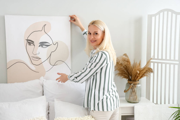 Mature woman hanging painting on light wall in bedroom - Foto, Bild