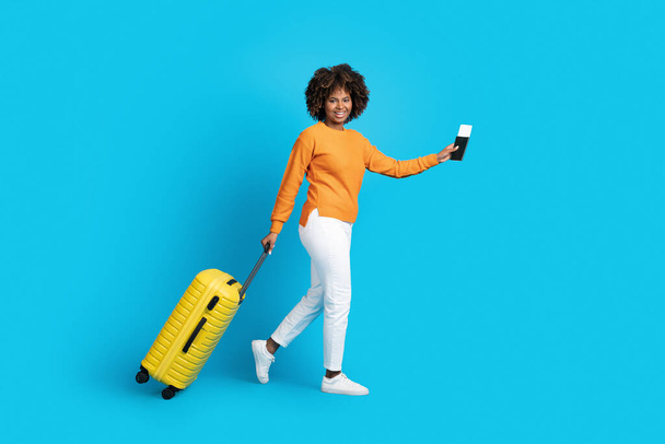 Šťastně se usmívající hezká mladá černoška cestující se žlutými zavazadly, s pasem a letenkami, modré studio pozadí, chůze směrem ke kopírovacímu prostoru, plná délka. Cestovní ruch, cestování - Fotografie, Obrázek