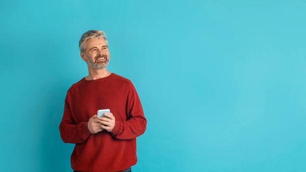 笑顔ハンサムな白髪の成熟した男は、彼の手の中にスマートフォンで青いスタジオの背景にポーズをとって、コピースペースを見て、素敵なモバイルアプリをチェックして、パノラマ - 写真・画像