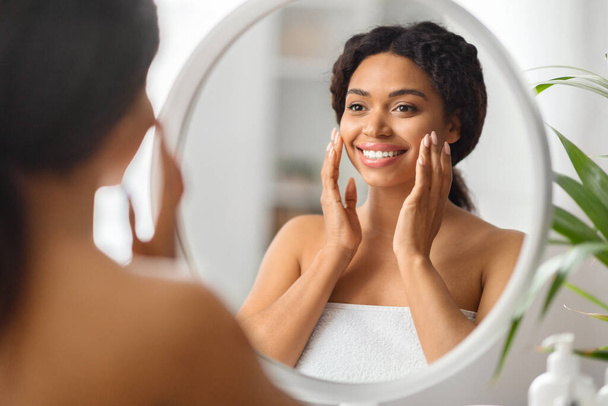 Schoonheidsconcept. Aantrekkelijke jonge zwarte vrouw op zoek naar spiegel in de badkamer, mooie Afro-Amerikaanse vrouw aanraken van haar gezicht en glimlachen tot reflectie, Genieten van huidverzorging Routine, Selectieve Focus - Foto, afbeelding