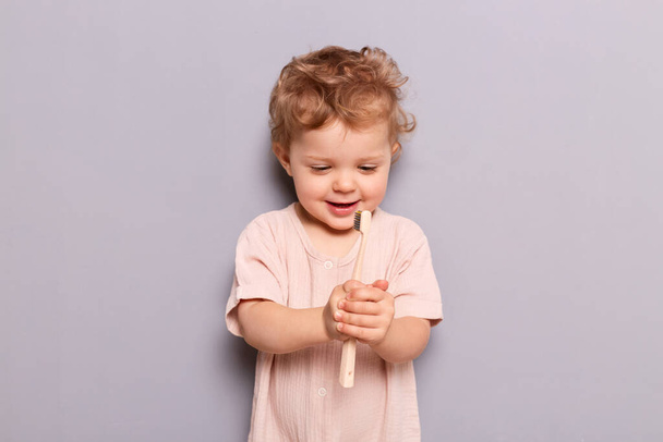 Cute little child baby brushing teeth isolated over gray background holdong looking at toothbrush with curious happy face learning taking care of her teeth by herself. - Photo, image