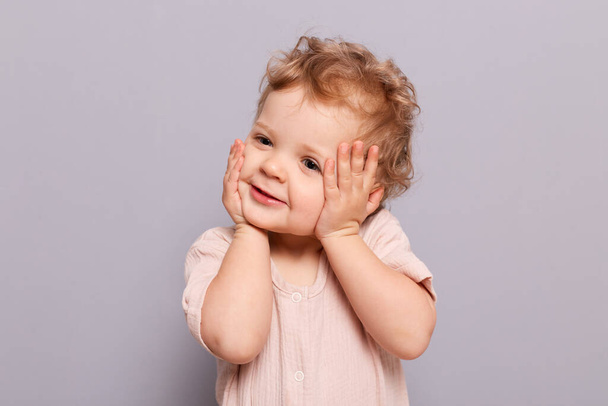 Cute charming smiling infant baby little caucasian girl posing isolated over gray background looking away with smile touching her face sees something interesting. - Photo, Image