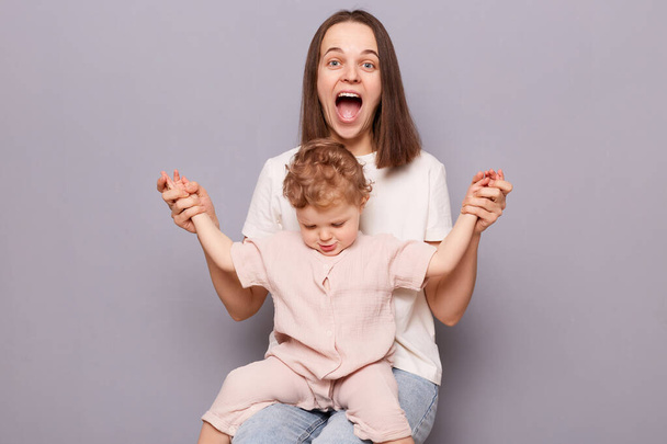 Mother and Baby. Happy woman with toddler child sitting sitting together isolated over gray background, mommy holding kid's hands and screaming with happiness and joy. - Foto, Bild