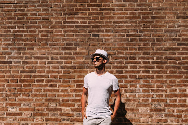 Ένας τύπος με γενειάδα με άσπρα ρούχα και γυαλιά ηλίου κρατάει τα χέρια στις τσέπες κοντά στον τοίχο από τούβλα. - Φωτογραφία, εικόνα