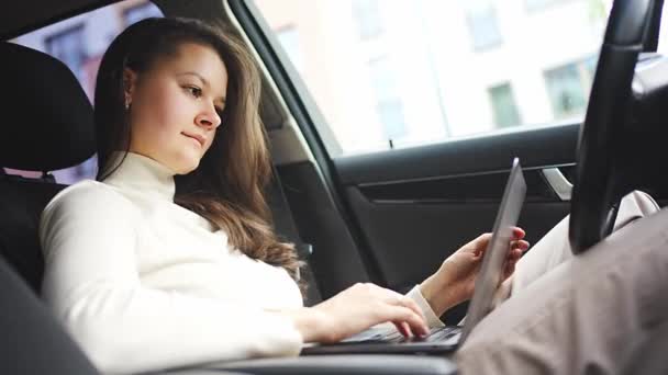 Νεαρή επιχειρηματίας που εργάζεται σε φορητό υπολογιστή κάθεται σε ένα αυτοκίνητο πίσω από το τιμόνι, χώρο εργασίας αυτοκινήτων. Υψηλής ποιότητας 4k πλάνα - Πλάνα, βίντεο