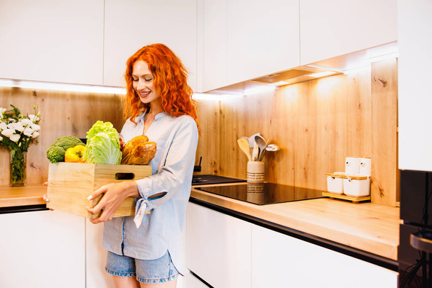 Happy healthy young woman with fresh vegetables in a green plastic box in her arms standing grinning at the camera in their kitchen. High quality photo - Photo, Image