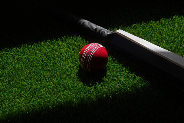 Murciélago de cricket y bola roja con iluminación natural sobre hierba verde. Cartel de tema deportivo horizontal, tarjetas de felicitación, encabezados, sitio web y aplicación - Foto, Imagen