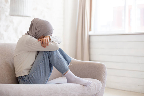 Young Muslim female student in hijab sitting on sofa at home, resting head on knees and crying. Problems with studies, parents, boyfriend. - Foto, Bild