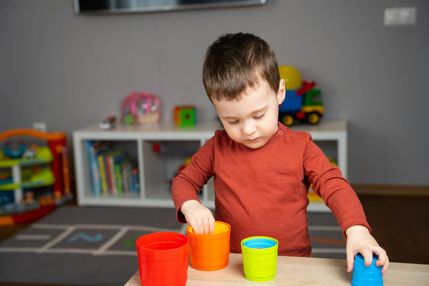 A cute little toddler boy of two years old sits at a children's table and plays with multi-colored pyramids in his room. Educational toys for shildren. Soft selective focus - Фото, зображення