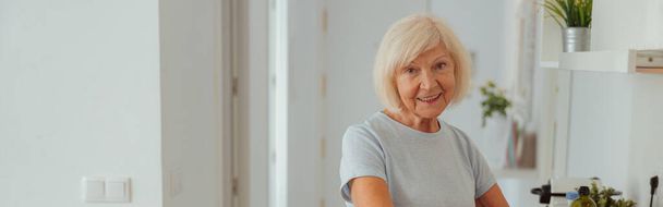 Foto de mulher idosa alegre vestindo roupas casuais sorrindo enquanto prepara o café da manhã na cozinha moderna em casa - Foto, Imagem