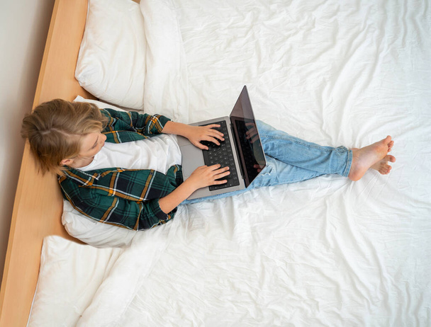 Teenage child boy using laptop. Child typing on the keyboard lying in bed. Play video game. Schoolboy studying doing homework. E-learning, remote educational classes top view - 写真・画像