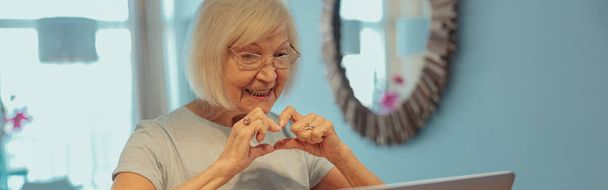 Sorridente vecchia femmina con i capelli grigi in occhiali che mostra il gesto del cuore durante la chiamata webcam sul computer portatile nella sua casa - Foto, immagini