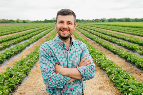 portrait of a modern bearded farmer man with crossed arms looking at camera stands in the agricultural field, Cheerful male worker in agricultural farm. Agriculture farming. - 写真・画像
