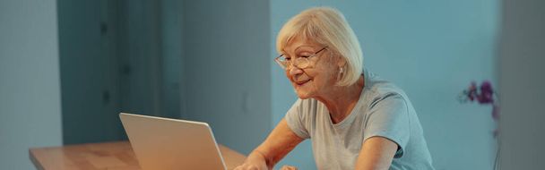 Donna anziana con i capelli grigi guardando lo schermo del computer portatile con espressione facciale felice quando si siede sulla scrivania del posto di lavoro a casa - Foto, immagini
