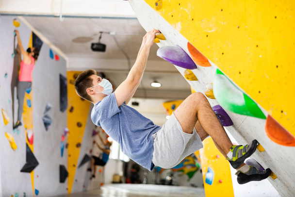 European young man in face mask grabbing ledges of artificial climbing wall in bouldering centre. - Фото, изображение
