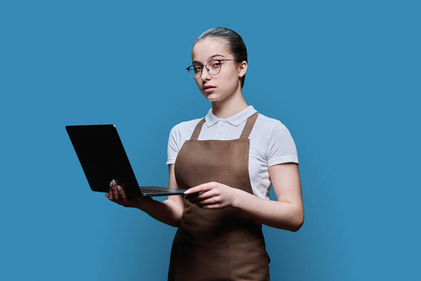 Young female student worker in apron using laptop, on blue studio background, teenage girl looking at camera. Youth employment in services, staff of shop cafe store - Foto, immagini