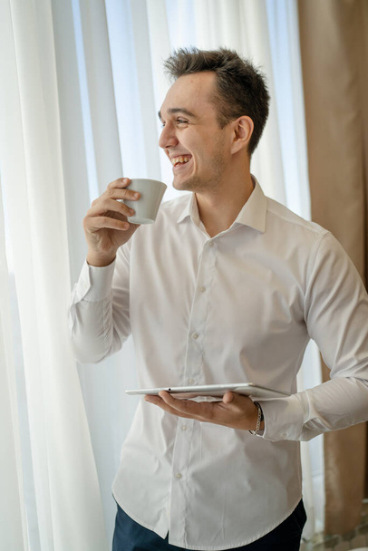 One man adult caucasian male standing by the window at home office or hotel room with cup of coffee and digital tablet taking a brake from work and preparing real people copy space wear white shirt - Photo, image