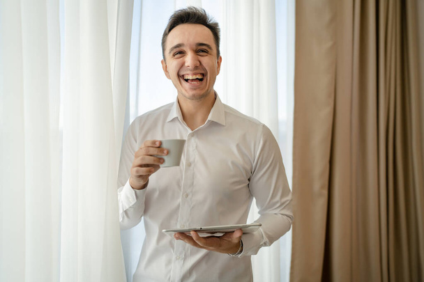 One man adult caucasian male standing by the window at home office or hotel room with cup of coffee and digital tablet taking a brake from work and preparing real people copy space wear white shirt - Foto, Imagen