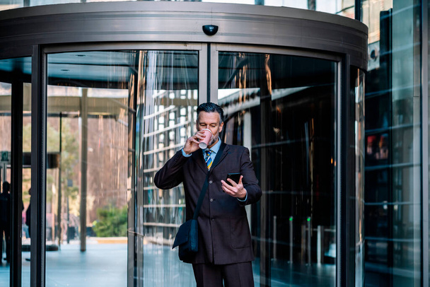 Pomyślny dorosły biznesmen w eleganckim garniturze przeglądający telefony komórkowe i pijący napoje na wynos stojąc przy wejściu do nowoczesnego budynku biurowego przy ulicy - Zdjęcie, obraz