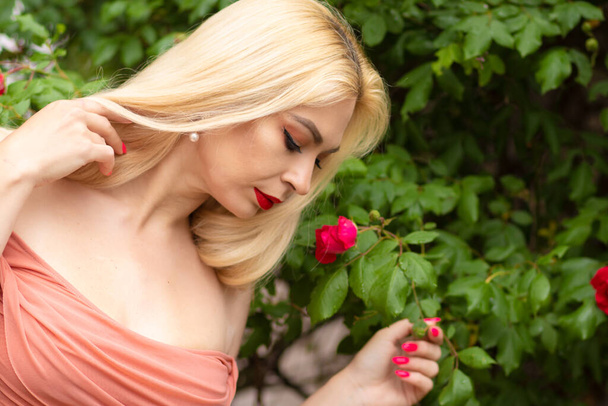 Beautiful woman with long blonde hair and red lips wearing pink clothes poses near blooming roses in garden. Wear light pink top and skirt - Фото, изображение