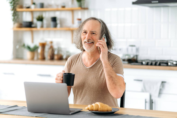 Zadzwoń do mnie. Pozytywny biały seniorek siwy biały mężczyzna, siedzi przy stole w domu w kuchni, używając laptopa, rozmawia przez smartfona z przyjacielem, pije kawę, uśmiecha się szczęśliwie - Zdjęcie, obraz