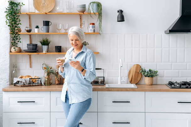 Великолепная кавказская седовласая женщина средних лет, стоит дома на кухне, держит смартфон и стакан кофе, общается в социальных сетях, читает новости, делает онлайн заказ, улыбается - Фото, изображение