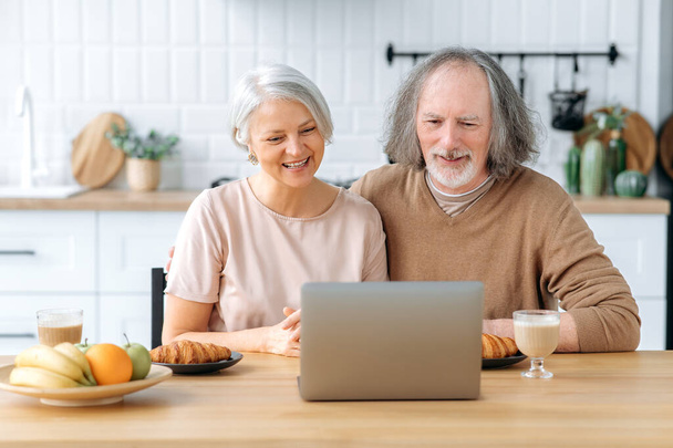 Positive caucasian pensioner couple, caucasian husband and wife, sit at home in the kitchen, have croissants and coffee for breakfast, use a laptop during breakfast, read news, watch videos, smile - Foto, afbeelding