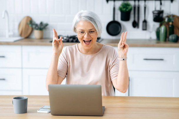 Joyful mature modern caucasian gray haired woman, with glasses, sits in the kitchen at a table with laptop, works remotely, manages finances, rejoices at result, smiles happily, gesturing hands - Foto, afbeelding