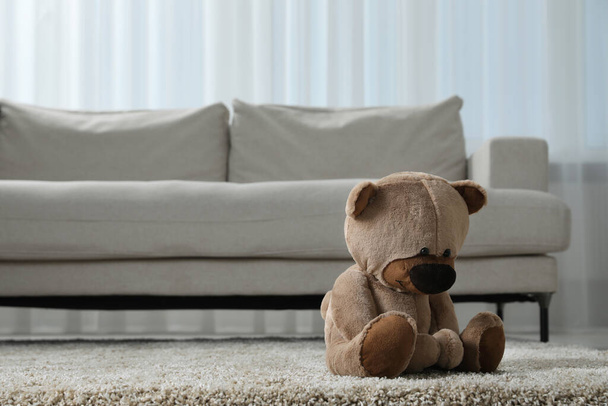 Cute lonely teddy bear on floor near sofa in room. Space for text - Foto, Imagen