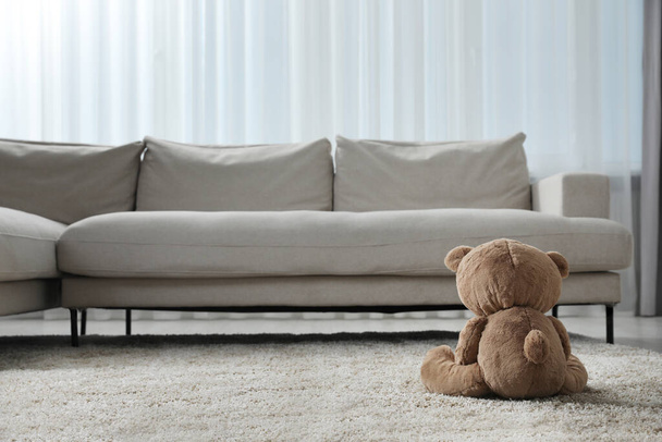 Bonito oso de peluche solitario en el suelo cerca del sofá en la habitación, vista trasera. Espacio para texto - Foto, Imagen