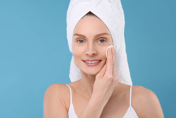 Beautiful woman in terry towel removing makeup with cotton pad on light blue background - 写真・画像