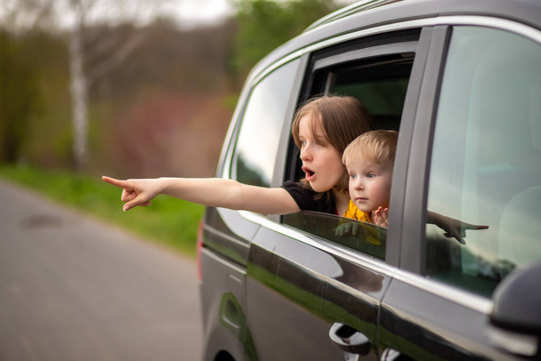 Mutlu erkek ve kız kardeş araba camından dışarı bakar, parmağını yana doğru tutar ve şaşırır. - Fotoğraf, Görsel