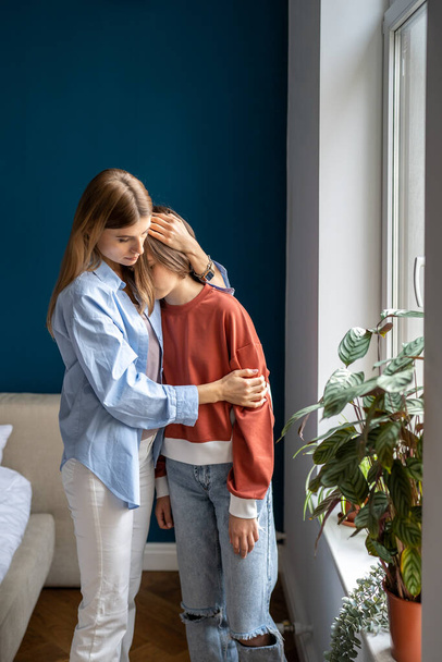Loving older sister embracing teen girl, parent single mother hugging supporting teenage daughter while standing together near window at home. Trust and understanding in parent child relationship - Photo, Image