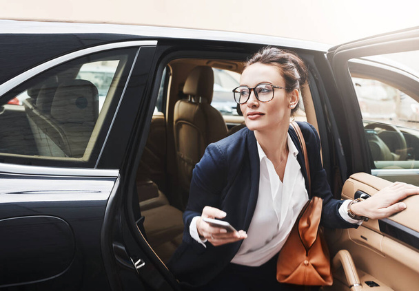 I have to hurry or Ill be late. a confident young businesswoman getting out of a car while holding a cellphone outside during the day - Фото, изображение