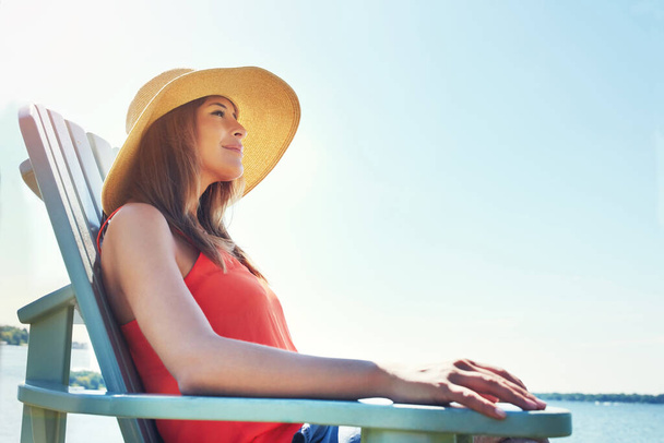 Ειρήνη και γαλήνη. Μια χαρούμενη νεαρή γυναίκα που φοράει καπέλο ενώ κάθεται σε μια καρέκλα δίπλα σε μια λίμνη έξω στον ήλιο - Φωτογραφία, εικόνα