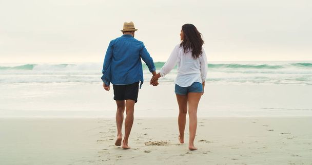 Wir lieben es, Zeit am Strand zu verbringen. Rückansicht in voller Länge von einem anhänglichen jungen Paar, das tagsüber nebeneinander am Strand spaziert - Foto, Bild